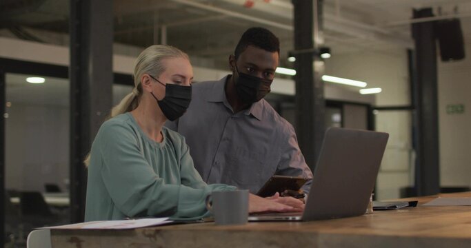Collaborating masked colleagues examining smartphone typing at office desk with laptop coffee mug