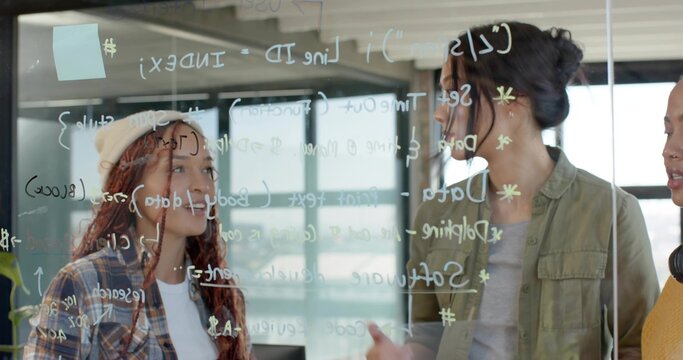 Women in varied attire collaborating on code behind glass board in office, with pastel sticky note