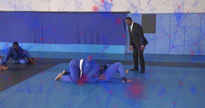 Grappling partners wearing blue gis, white belts on dojo padded mats, with coach in suit observing