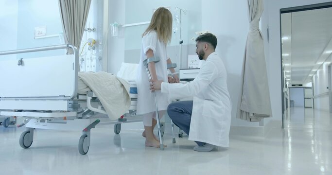 Kneeling doctor helping child in gown using crutches in ward, with bed, IV pole, cart, curtains - Powered by Adobe