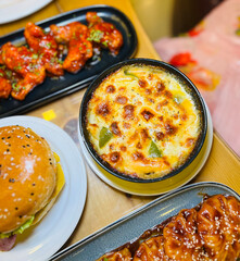 Close up of a delicious cheesy baked dish with a golden brown crust served alongside a juicy burger and spicy appetizers on a wooden table