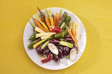 A variety of vibrant miniature vegetables, including carrots, eggplants, cucumbers, and baby corn, are arranged artfully on a white plate, set against a bright yellow background