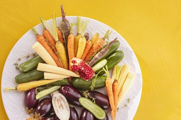 An array of fresh colorful vegetables are arranged on a white plate. Carrots, baby corn, zucchini, and eggplants create a healthy and visually appealing snack
