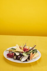 A White plate holds a colourful arrangement of fresh raw vegetables, including mini eggplants, baby carrots, cucumbers, and pomegranates, all on a bright yellow background