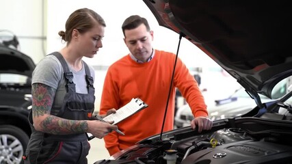 A female mechanic with tattooed arms holds a clipboard and discusses car repairs with a male customer standing next to an open car hood in a garage. - Powered by Adobe