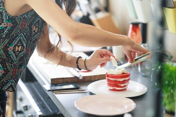 A person carefully layers fresh mozzarella onto slices of ripe red tomatoes, creating a visually appealing Caprese tower on a pristine white plate. A culinary artwork takes shape