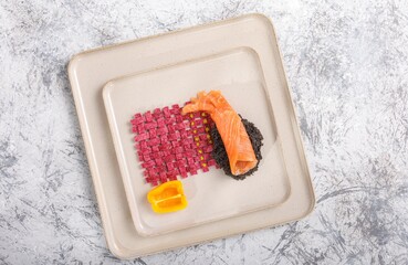 A delicate salmon roll rests on black rice, alongside a woven beetroot structure accented with yellow spheres, completed with a yellow pepper, all displayed on minimalist square dishware