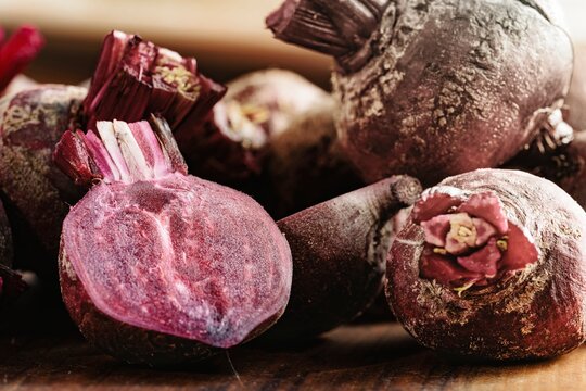 A collection of earthy beets rests on a dark wooden surface, some whole, others halved to reveal their deep purple interiors. The beets show fresh and organic textures, making a rustic scene