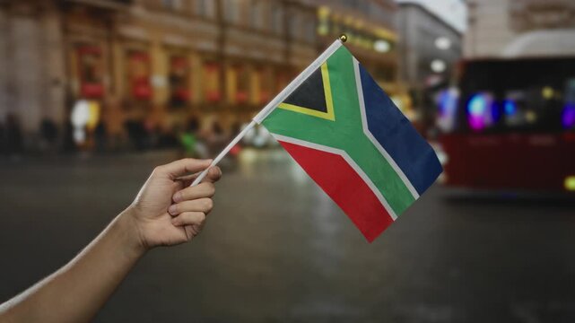 Hand holding south african flag in city street capturing dynamic cultural essence in urban setting during evening with diverse people and lights in background.
