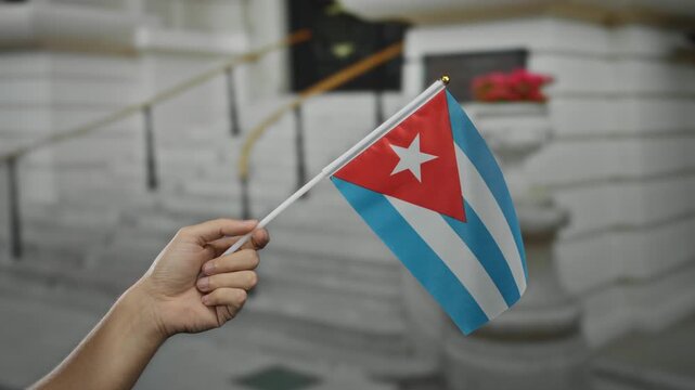 Caucasian man holding cuban flag outdoors in city street with focus on hand against blurred urban background showcasing national pride and identity.