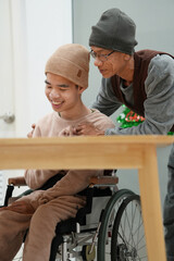 Young man with disability wearing winter pajamas on wheelchair with his father or caretaker in the house with a christmas tree decoration,Winter family activities,Happy New Year in sweet home concept.