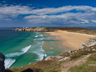 Turquoise waves sweep a wide golden beach beneath rugged cliffs at Pointe de Pen Hir on the Crozon Peninsula, Brittany, France, under a bright blue sky.