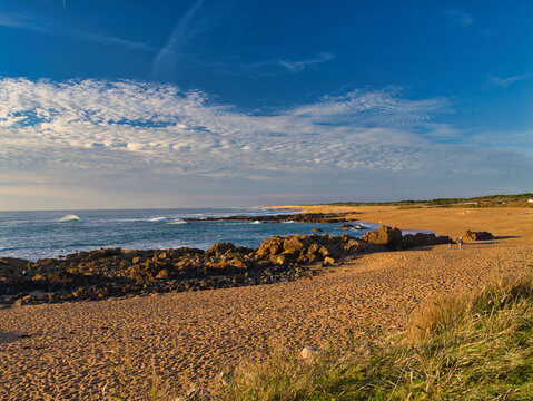 Warm sunset light bathes a wide sandy shore and rocky outcrops at Les Sables-dOlonne on the Vendee coast, France, with gentle waves and patterned clouds.