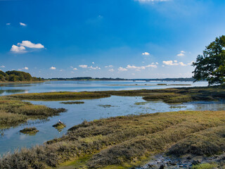 Wide view of tidal wetlands and calm water at Ile de Conleau near Vannes, Brittany, France, under a blue sky with scattered clouds and distant boats