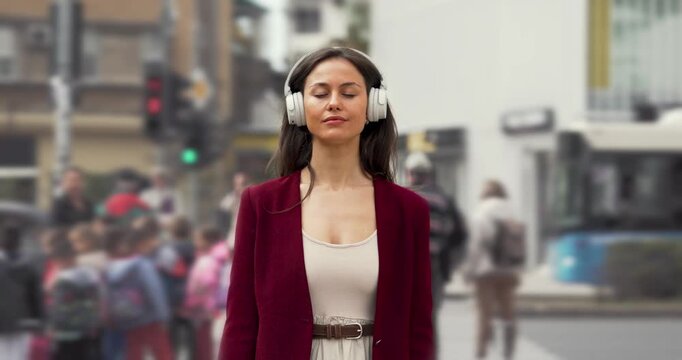 Beautiful young woman meditating with closed eyes and headphones in busy urban street setting, wearing burgundy business attire, time lapse.