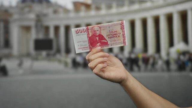Male hand holding icelandic krona banknote in an outdoor vatican city setting with blurred background of san pedro square architecture.