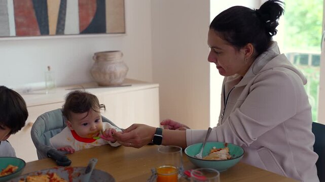 Mother feeding baby at dining table, nurturing family moment showing care, patience, and everyday love during shared mealtime