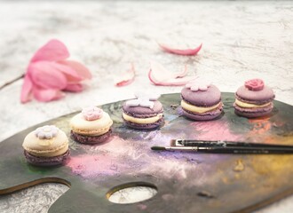 A painterly arrangement shows delicate macarons topped with flower accents resting on a colorful artist's palette with brushes. A pink flower and petals complete the artistic tableau