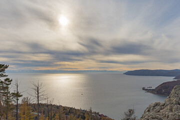 Sunlit coastal panorama over Baikal Lake with cliffside Rocks, distant Khamar-Daban Mountains and autumn forests of larch and fir in Siberia, Russia.