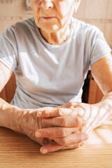 An elderly woman sits at a table in a sunlit room, her hands folded on the table, close-up of her hands