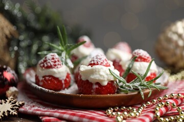 Christmas food. Santa hats made of strawberries, whipped cream, rosemary and decor on table against background with blurred lights, closeup. Bokeh effect