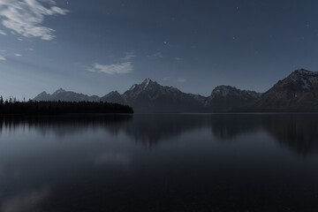 Moonlit Colter Bay Beach with still lake and Teton mountains under night sky.