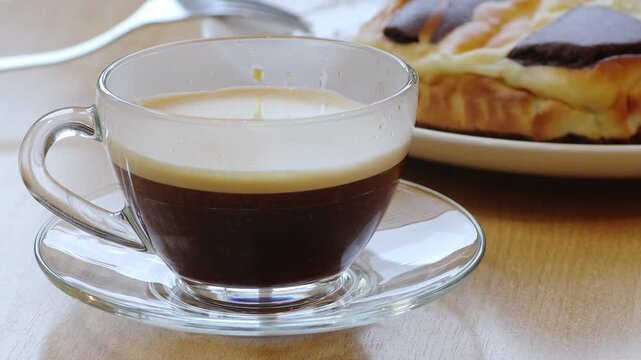 Closeup video of a hand placing a transparent glass cup of coffee on a wooden table and then pouring fresh milk into it.