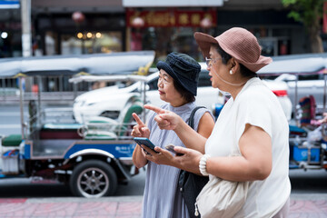 Two senior asian women travelers holding smartphone and pointing while discussing direction beside tuk tuk showing friendship and city travel lifestyle happiness and active retirement tourism