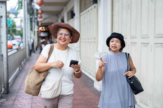 Senior asian women travelers smiling walking along city sidewalk holding smartphone talking and pointing showing friendship freedom curiosity and joyful retirement lifestyle