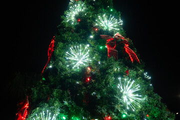 A tall evergreen Christmas tree glows with red, green, and white LED starburst lights. Big bows and ornaments shine brightly against a dark night sky.