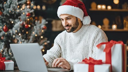 Smiling man in Santa hat and white sweater using laptop for Christmas online shopping or remote work with gifts and decorated tree. Holiday tech usage, gift preparation