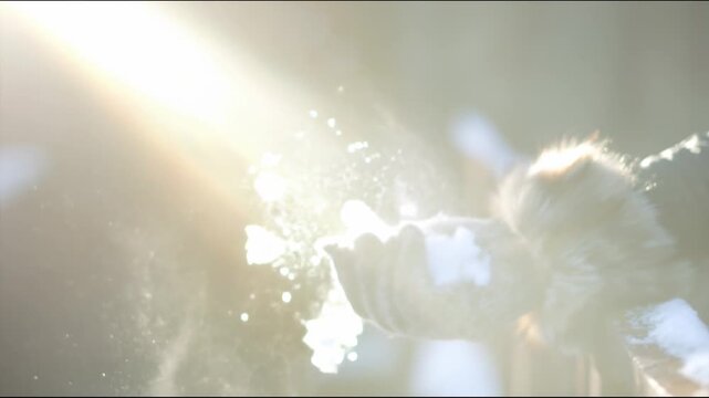 A hand in a gray knitted mitten with fur trim holds snow that scatters into the air under the winter sunlight