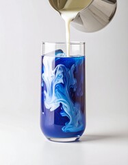 A clear glass is filled with a vibrant blue latte, with a stream of milk being poured into it from a metal pitcher, creating swirling patterns. Blue latte with butterfly pea flower tea. 
