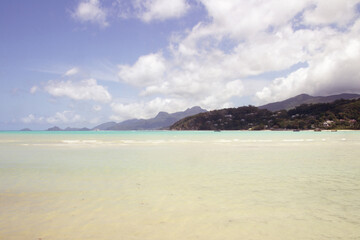 Shallow turquoise lagoon of Mahé Island in Seychelles, with distant mountains and tropical coastline under bright sunlight and scattered clouds.