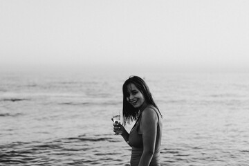 A cheerful woman in a vibrant red swimsuit stands by the seaside, holding a glass of white wine. The setting sun casts a warm golden hue over the scene, highlighting her joyous expression.