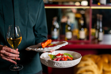 A person holds two plates of fine dining food and a glass of white wine in a restaurant setting....