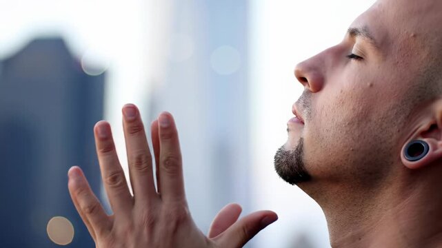 Extreme close-up of a mid-30s man with a goatee and ear gauges, eyes closed, hands praying then gesturing upwards. Blurred urban background. Concept of spiritual devotion