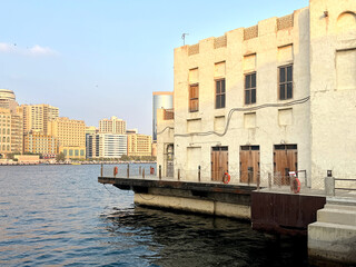 Traditional waterfront building with wooden doors on Dubai Creek, contrasting with modern city skyline at sunset.