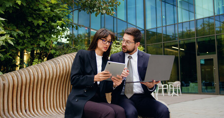 Two business partners man and woman in formal attire sitting on bench outside a modern office building, discussing work-related topics using a digital tablet and laptop.