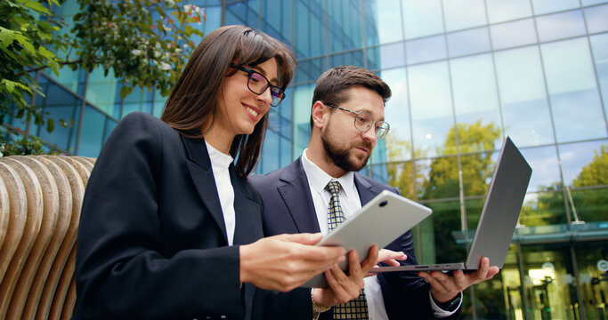 Side view of two successful colleagues man and woman having outdoor meeting, discussing important work topics using laptop and tablet near office building