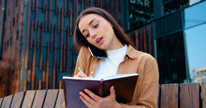Close up of self employed female making phone call on mobile phone and taking notes while working remotely while sitting on bench about modern office building, outdoors