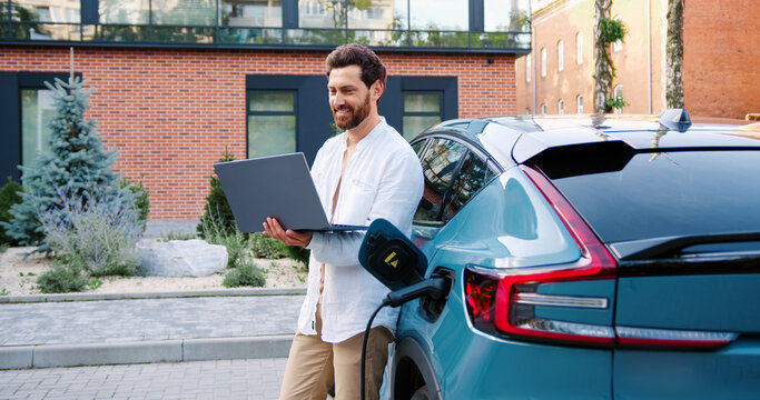 A handsome man uses a laptop app while standing next to his electric car charging at portable station on city street, with urban buildings in the background.