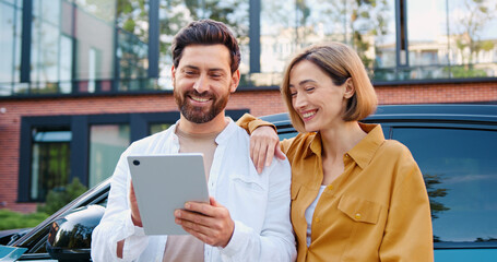 Portrait of happy young couple using a tablet outdoors, talking and smiling, with a car and modern cityscape behind them