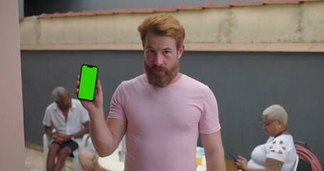 Man holding smartphone with green screen chroma key, standing outdoors in courtyard, presenting phone to camera, casual social setting with seated people in background