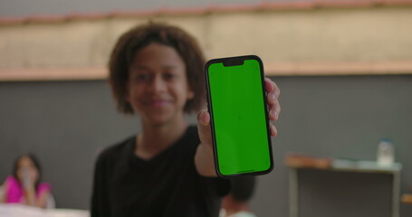 Teenager of African descent holding smartphone with green screen, chroma key feature displayed, smiling outdoors at a casual gathering with diverse adults in the background
