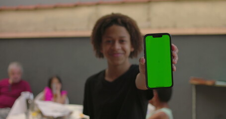 Teenager of African descent holding smartphone with green screen, chroma key feature displayed, smiling outdoors at a casual gathering with diverse adults in the background