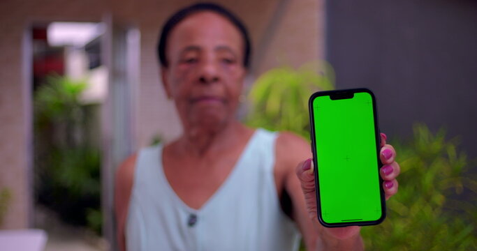 Elderly African American woman holding smartphone forward, green screen Chroma Key on display, blurred face in background, concept of digital technology and communication outdoors