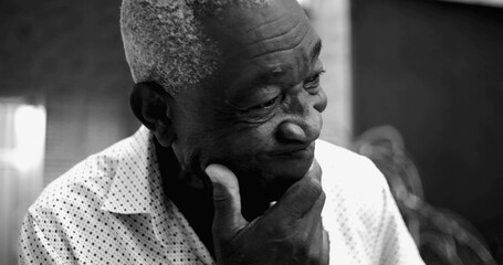 Elderly Afro-descendant man in black and white portrait, eyes closed with a pensive and slightly concerned expression, hand resting on chin in a reflective home setting