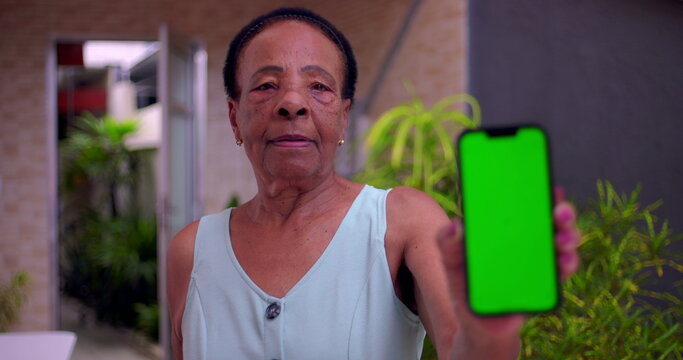Elderly African American woman holding smartphone forward, green screen Chroma Key on display, blurred face in background, concept of digital technology and communication outdoors - Powered by Adobe