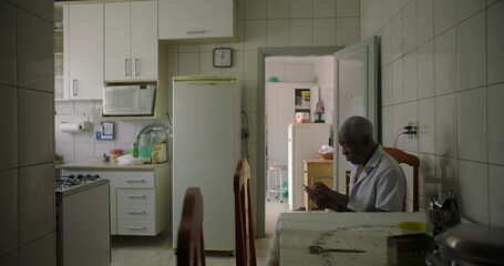 One Elderly Black man sitting at dining table in a cozy kitchen, concentrating on a smartphone, capturing the essence of daily routine and technology in a relaxed home environment